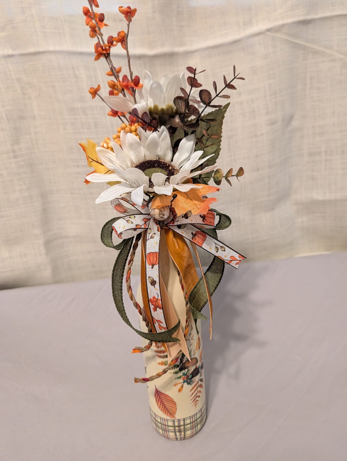 Decoupaged bottle in cream and plaid, filled with white daisies, orange maple leaves and trailing ribbon
