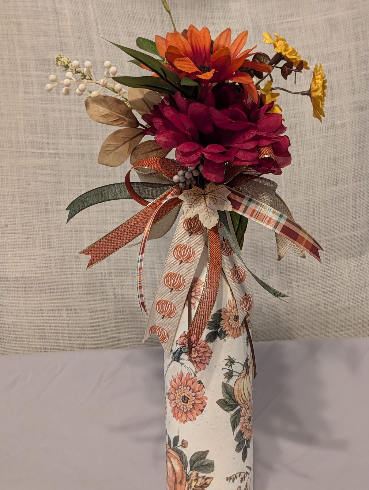 Decoupaged bottle in a pumpkin and mum print, topped with a burgundy dahlia, orange gerbera and ribbon spray