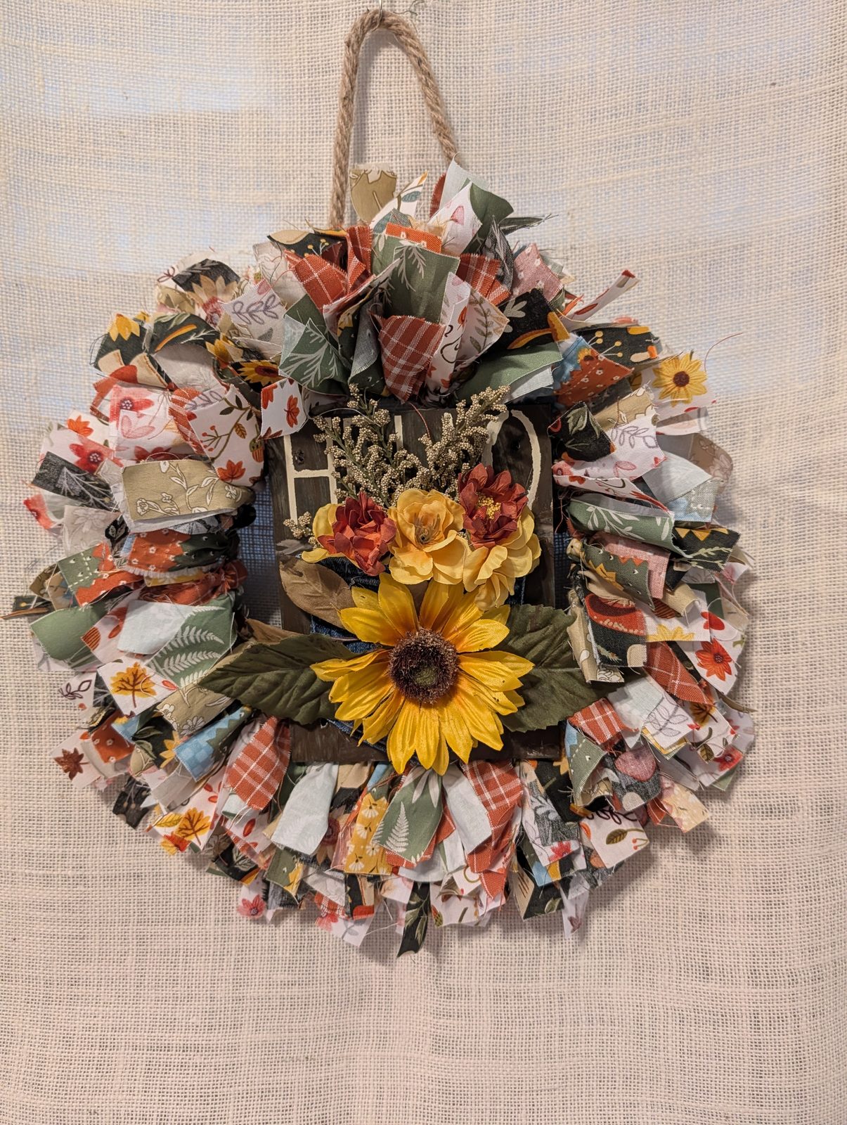 Rag wreath tied from autumn print fabrics around a wood plaque, finished with a sunflower and cream blooms