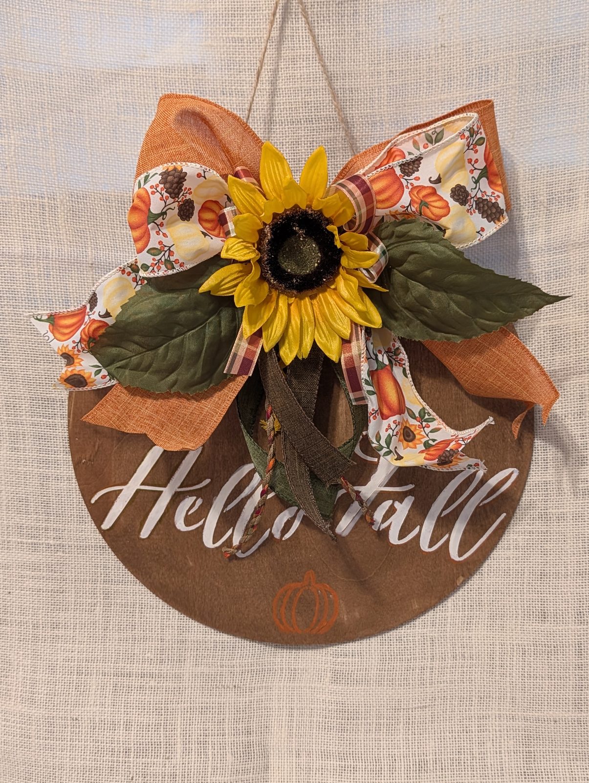Round stained-wood door hanger lettered &lsquo;Hello Fall&rsquo; with a sunflower and pumpkin-print bow