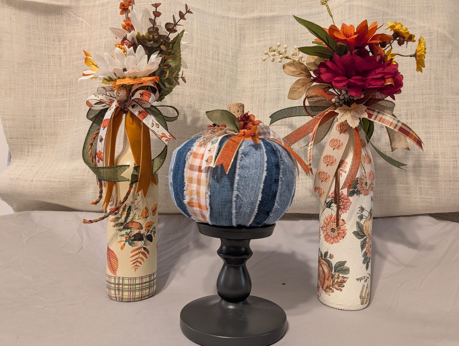 Two decoupaged bottles with autumn flowers flanking a denim patchwork pumpkin on a stand